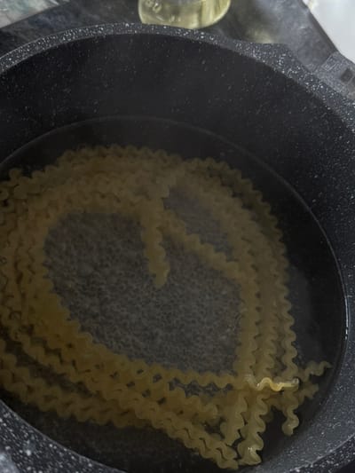 The image displays an overhead view of a dark grey cooking pot, potentially featuring a speckled non-stick coating on its upper rim. Inside the pot, water is actively boiling, evidenced by numerous small bubbles concentrated across its surface. Partially submerged in the boiling water are multiple strands of light yellow pasta. The pasta is characterized by its long, flat, ribbon-like form with distinct ruffled or wavy edges, consistent with varieties such as Mafaldine. No individuals are present in the frame. In the blurred upper right background, a translucent yellow object, possibly a bottle, is visible resting on a dark, indistinct surface, likely a kitchen counter or stovetop. The scene is purportedly located in Bad Nauheim, Germany.