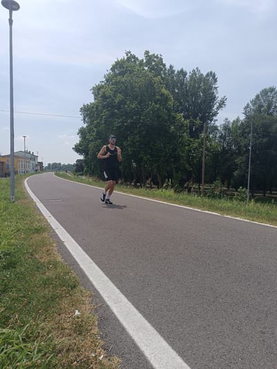 A man is running on a paved road in Casalmaggiore, Italy. He is wearing a black tank top, black shorts, white socks, and black and white running shoes. He has sunglasses on his head and is wearing a watch. The road has a white line on the left side of it. There is grass and trees on both sides of the road. In the background, there are buildings and more trees. The sky is cloudy.
