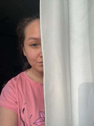 A medium shot depicts a person partially visible from the chest up, peering from behind a white fabric, presumably a curtain or sheet. The person has fair skin, light brown hair, and one visible eye that is brown. They are wearing a pink t-shirt with a dark floral or abstract design on the upper right. The background is dark, appearing to be an interior space, possibly a dimly lit room. The photograph is oriented upside down. The image shows no discernible evidence of a location in Verkh-Katunskoye, Russia.
