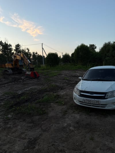 An excavator and a white car are parked on a muddy construction site in Akul'shino, Russia. The excavator, a yellow Hyundai, is positioned on the left side of the image. It has a red and black attachment on its bucket. The car, a white sedan, is on the right side of the image and appears dirty. The ground is covered in mud and some patches of grass. In the background, there are trees and a power line pole. The sky is light blue with some wispy clouds, suggesting it is either early morning or late evening. There are no people visible in the image.