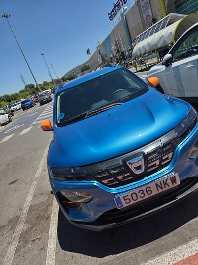 In the city of Collado Villalba, Spain, a bright blue Dacia Spring electric vehicle is parked in a parking lot adjacent to a Carrefour supermarket. The car's license plate reads "5036 NKW". A white vehicle is parked to the right of the blue car, partially obscuring it. In the background, the Carrefour building is visible with its signage, along with streetlights and trees. The sky is clear and blue, indicating a sunny day. The parking lot has lane markings and some other vehicles can be seen in the distance.