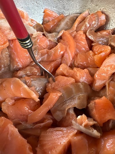 A close-up, overhead shot captures a bowl filled with bite-sized pieces of raw salmon. The salmon, cut into irregular shapes, is a vibrant orange-pink color and appears to be lightly seasoned with black pepper. Some pieces have a slightly lighter, almost translucent quality, while others are more opaque. A red-handled fork with silver tines is partially submerged in the salmon, with its tines gripping a few of the pieces, suggesting the salmon is being prepared or mixed. The background appears to be a silver or metallic bowl, reflecting some of the light. There is no discernible information about a location in Zheleznodorozhnyi, Russia.