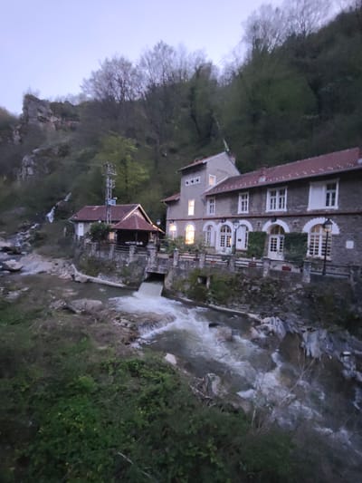 The image shows a building complex near a river or stream, likely at dusk. The main building is a multi-story stone structure with a red-tiled roof and arched windows. A smaller, adjacent building has a similar red-tiled roof and a covered porch. Both buildings are situated close to a body of water with rapids. The location is Vucje, Serbia. The surrounding environment includes a steep, forested hill. The illumination suggests the buildings have internal lighting, with some windows glowing. The water flows downstream from what appears to be a culvert or opening beneath the building, suggesting a historical or current water mill setup. Vegetation is present in the foreground along the banks of the waterway. No people are visible.
