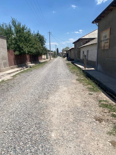 The image depicts a street in the city of Боғистон, Tajikistan, on a clear day. The street is unpaved, covered with gravel and dirt. On the left side of the street, there is a tall brown metal fence backed by a dense row of green trees. A gray concrete wall extends from the fence. On the right side of the street, there are several buildings with sloping roofs. One building has a sign with Cyrillic text that reads "УЗИ ОПОЛЮГИЧЕСКИ СОЛЯРИЙ". A narrow concrete drainage ditch runs along the edge of the sidewalk next to the buildings. The sky is bright blue with a few scattered clouds. There are utility poles with electrical wires running along the street. In the distance, the street curves and continues into the city. There are no people clearly visible in the foreground, though there might be figures further down the road. The scene suggests a residential or semi-rural area within the city.