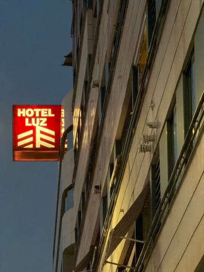 The image shows a close-up of a red sign for "HOTEL LUZ" with white text and a stylized white logo. The sign is illuminated and appears to be attached to the side of a building. To the right of the sign is a multi-story building with a facade composed of diagonal white panels and dark vertical windows. The lighting suggests it is dusk or night. The building's architecture is modern with visible structural elements and some external fixtures. The sky in the background is a muted blue. The sign is positioned on the left side of the frame, while the building occupies the majority of the right side.