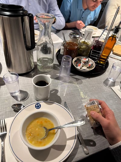A top-down perspective captures a breakfast scene. A man in a blue shirt sits to the left. Various breakfast items are arrayed across the table. There's a rack holding multiple jars of condiments, including what appears to be honey, jam, and pickles. A bowl containing individual creamers sits nearby.
A person's hand on the right holds a small package of bread. A bowl of soup with a spoon sits on a plate in the lower right. A coffee mug filled with dark liquid and a glass water carafe are also present. A metal coffee server sits on the bottom left. Cutlery is arranged around the soup bowl. The location is unknown and cannot be ascertained from this image. The city of Mirabel, Canada is also unable to be discerned from this image.