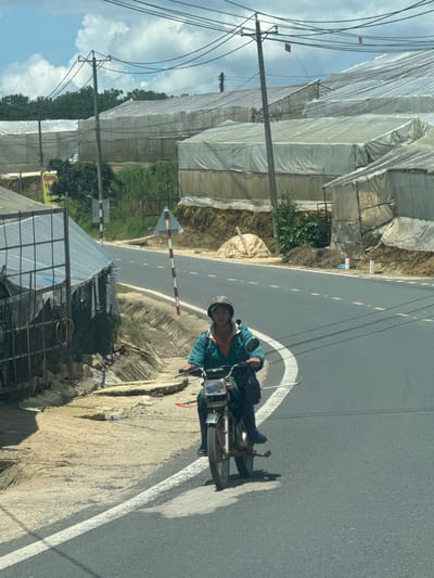 A person wearing a green jacket and a helmet is riding a motorcycle on a paved road in what appears to be a rural or semi-rural area. The person is looking towards the camera. The road has yellow dashed lines indicating lanes. On the left side of the road, there is a structure with a metal fence and a translucent roof, and further back, a hillside with greenhouses covered in white plastic. On the right side of the road, more greenhouses are visible, as well as power lines and utility poles. The sky is partly cloudy. A yellow sign with red text is visible on the left side, partially obscured by trees.