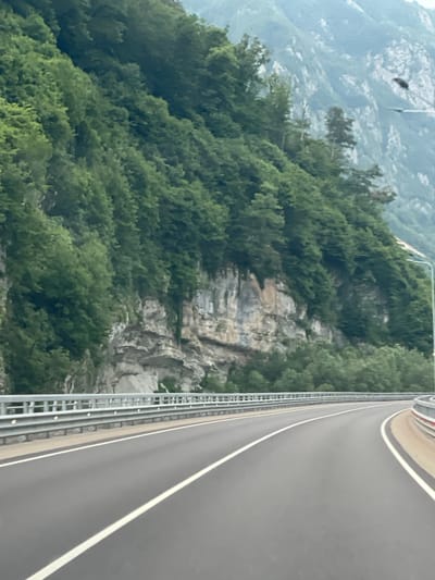 The image depicts a curving asphalt road with white lane markings, bordered by a metal guardrail on the left and a sandy shoulder with a red-and-white barrier on the right. To the left of the road, a steep, tree-covered mountainside rises dramatically, with exposed rock faces visible in sections. In the distance, further mountains are shrouded in a hazy, overcast sky. A streetlamp is visible on the right side of the road, positioned on the mountainside. No people are present in the image. The scene suggests a natural, mountainous environment, likely a highway passing through a scenic area. The description is not specific to the city of Monastyr, Russia, as no identifiable landmarks or contextual clues place it there.