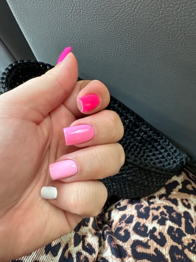 A close-up, overhead shot features a person's left hand with a manicure of different colored nails against a dark gray, textured surface and a black, woven material. The nails are painted in shades of pink, with the thumb nail a bright neon pink, the index finger a hot pink, the middle finger a lighter pink, and the ring finger a pale pink. The little finger is painted a light, muted gray. The hand is positioned as if holding or interacting with the black woven material, which appears to be part of a bag or accessory. Behind the hand and woven material, the dark gray surface has a distinct leather-like texture. The lower right portion of the image shows a piece of fabric with a leopard print pattern in shades of brown and black. The scene appears to be indoors, possibly in a vehicle or seated area, given the textures and lighting. There are no visible landmarks or context to suggest a specific location within Мастоцкі сельскі Савет, Belarus.