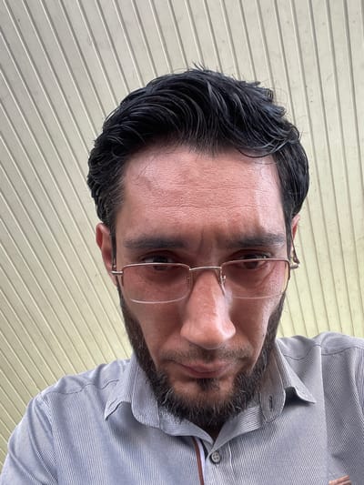 A man with dark hair, a beard, and glasses is looking down at the camera. He is wearing a light blue striped shirt. The background appears to be an outdoor ceiling with parallel lines. The location is Mashrab, Uzbekistan.