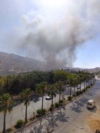 This media file depicts a dense plume of dark smoke rising from an urban area, likely a residential district, in Nablus, Palestinian Territories. The smoke billows into a clear blue sky, contrasting sharply with the bright sunlight, suggesting a daytime event.
The scene is dominated by the large smoke cloud, which obscures the background buildings and hillsides. The urban landscape features a dense collection of multi-story buildings cascading down a slope, characteristic of hilly cities. In the foreground, a paved road lined with palm trees runs parallel to the viewer. Several vehicles, including cars and a white van, are visible on the road, indicating normal traffic flow despite the smoky atmosphere.
The presence of palm trees suggests a warm climate. The overall impression is one of an active event, possibly a fire or explosion, unfolding within a densely populated city. The thick smoke implies a significant incident. The image conveys a sense of immediate concern due to the visible hazard. There is no discernible text in the image.