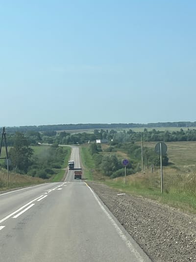 The image shows a rural road in the city of Урталга, Russia, on a clear, sunny day. The road is paved and has double white lines marking the lanes. Two trucks are visible on the road, one in front of the other, traveling uphill. The landscape on either side of the road is green and rolling, with trees and fields. There are also power lines and a few signs visible along the roadside. The overall impression is one of a quiet, rural setting on a summer day.