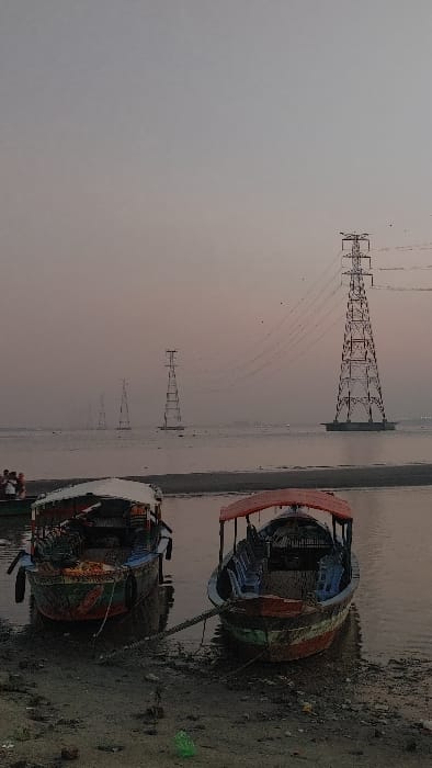 This image depicts a waterfront scene at dusk or dawn in Kumarvog, Bangladesh. In the foreground, two traditional wooden boats with canopies are partially pulled onto a muddy and sandy shore. The boat on the left has a white canopy, a blue hull with green and red accents, and an apparent outboard motor mounted at the stern. The boat on the right features a red canopy and contains multiple light blue plastic chairs arranged within its interior. A thick rope extends from this boat towards the lower right, disappearing out of frame. The immediate shoreline is littered with small rocks, detritus, and a bright green plastic bottle.

In the midground, a body of water, likely a river or lake, stretches horizontally across the frame. On the far left, a small cluster of indistinct individuals stands near the water's edge, and other small boats are partially visible further out on the water to the left. The background is dominated by a series of large metal lattice transmission towers (electricity pylons) extending across the water. The most prominent pylon, positioned on the right, stands on a concrete foundation in the water and supports multiple overhead power lines. Several other similar pylons recede into the hazy distance towards the left, gradually diminishing in size and clarity. The sky exhibits a gradient of muted grey-blue to a faint orange-pink hue near the horizon, indicative of low light conditions. Small dark specks, possibly birds, are visible in the sky around the largest pylon.
