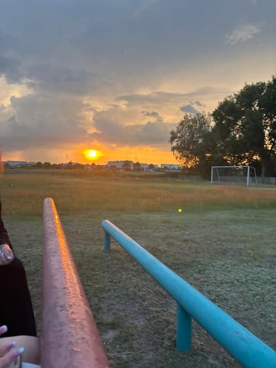 The image shows a sunset over a grassy field in the city of Сароцкі сельскі Савет, Belarus. The sun is setting behind a cluster of buildings, casting a warm orange glow across the sky and the landscape. In the foreground, two metal bars, one pink and one blue, are visible. A person's hand, with manicured nails, is visible near the pink bar. In the background, there is a soccer goal and a few trees. The overall scene depicts a peaceful, outdoor setting during twilight.