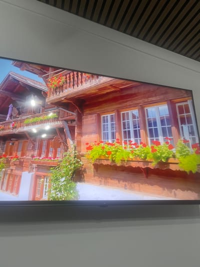 This is a photograph of a traditional wooden house, likely in a village or rural setting, with a clear blue sky and lush greenery visible. The house features multiple stories with balconies adorned with abundant red flowers in planters. The windows are also decorated with flower boxes filled with red blossoms and vibrant green foliage. The architecture suggests a European alpine style. The photograph is tilted slightly to the right. The location in the city of Rosenovo, Bulgaria is not discernible from the image itself.