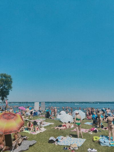 The image depicts a crowded beach scene on a sunny day in Nieporęt, Poland. The foreground is a grassy area with numerous people sunbathing on towels and blankets, interspersed with beach umbrellas and chairs. Several individuals are seen relaxing, some lying down and others sitting. In the mid-ground, the beach transitions to the water, which is filled with many people swimming and wading. Sailboats and other watercraft are visible on the lake. The background shows a hazy shoreline with trees and more distant structures. The sky is clear and bright blue.