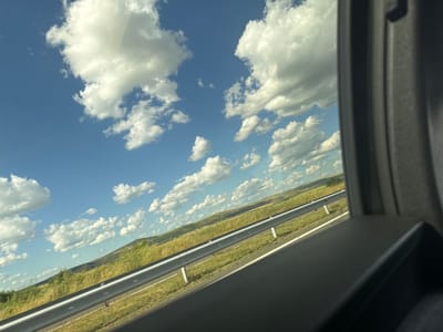 The image is a photograph taken from inside a vehicle, looking out at a landscape under a cloudy blue sky. The foreground shows a dark, textured surface that is part of the vehicle's interior, likely a window frame or dashboard. To the left of this, a metal guardrail runs diagonally across the frame, separating the road from the surrounding terrain. Beyond the guardrail, a grassy hillside slopes upwards, dotted with patches of yellow and green vegetation. The sky is a bright blue, filled with numerous fluffy white clouds, some of which are quite large and prominent. The overall impression is of a scenic drive through a rural area. The location is identified as Zheleznik, Bulgaria.