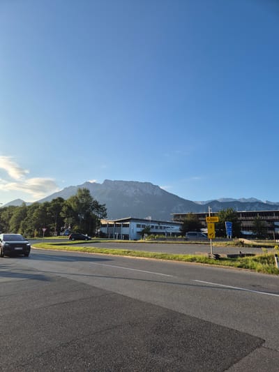 The image captures a scenic view of a mountain range under a clear blue sky, likely taken in Niederndorf, Austria, given the presence of a sign pointing to "Kufstein." In the foreground, a dark-colored car, identified by its license plate "EL 73 988," is driving on an asphalt road. The road curves to the left, with a grassy median strip separating it from another lane.
Behind the road, a modern building with a flat roof and multiple windows is visible. Adjacent to this building is a cluster of trees, with a taller mountain range forming the backdrop. A road sign with a yellow background and a symbol indicating a roundabout is positioned on the right side of the road. Another car is seen in the distance, also on the road. The overall scene suggests a clear day with good visibility, showcasing the juxtaposition of urban infrastructure and natural landscape.