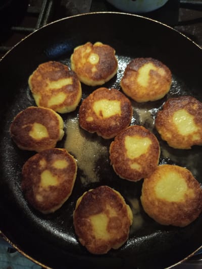 Ten syrniki, small, round, and golden-brown, are being fried in a black skillet. They are arranged in a circular pattern within the pan, with some of them overlapping slightly. The syrniki appear to be made from a mixture that includes cottage cheese and flour, as evidenced by their light yellow interior and golden-brown, slightly crispy exterior. Some of the syrniki have visible lighter patches in their center, suggesting uneven cooking or a characteristic of the batter. The skillet is on a dark, possibly gas, stovetop, and there is a small amount of melted butter or oil in the pan, giving the syrniki a slightly glistening appearance. The background is dark and out of focus, with a hint of a white object, possibly a bowl or plate, in the upper right corner. The overall impression is that of a home-cooked meal being prepared. The scene is indoors, likely in a kitchen. There are no people visible in the image. This image is not sufficient to determine the location in the city of Novotroitsk, Russia.