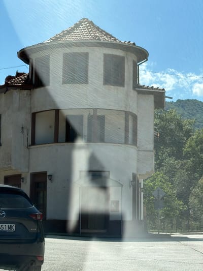 The image captures a slightly hazy view of a building with a curved facade and a tiled roof, situated in Beli Osam, Bulgaria. The building is a light color, possibly white or cream, with dark trim around the windows and doorways. It appears to have two stories. The windows on the upper level are square, while the ones on the lower curved section are arched. There is a street in the foreground with a dark SUV parked on the left side, its license plate reading "551 MK". Behind the building, lush green trees and a hillside are visible under a bright blue sky with some wispy clouds. The overall impression is a scene of a town or village setting, possibly on a sunny day, with the haze suggesting it could be warm or that the photo was taken through a window with some condensation or glare.