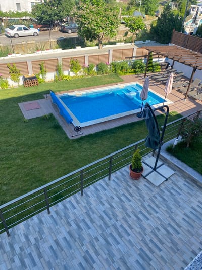 This is an aerial, high-angle, full shot of a backyard swimming pool and patio area in Ognyanovo, Bulgaria. The pool is rectangular with blue water and has a blue cover. There are lounge chairs and umbrellas near the pool. The patio is tiled and has a metal railing. A small green plant in a pot is on the patio. The backyard is surrounded by a fence and trees. In the background, there are houses and a street with cars. The overall scene depicts a residential area with outdoor recreational facilities.