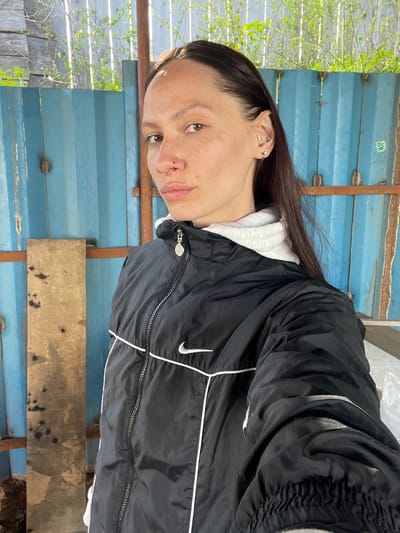 The image shows a woman with dark brown hair and fair skin wearing a black Nike jacket with white trim and a white hoodie underneath. She has multiple piercings in her left ear, a nose ring, and a labret piercing. She is standing outdoors in front of a structure made of blue metal sheets and wooden planks. Green foliage is visible on the left side of the frame. The location is assumed to be Doma 45 Kilometr, Russia based on user provided information.