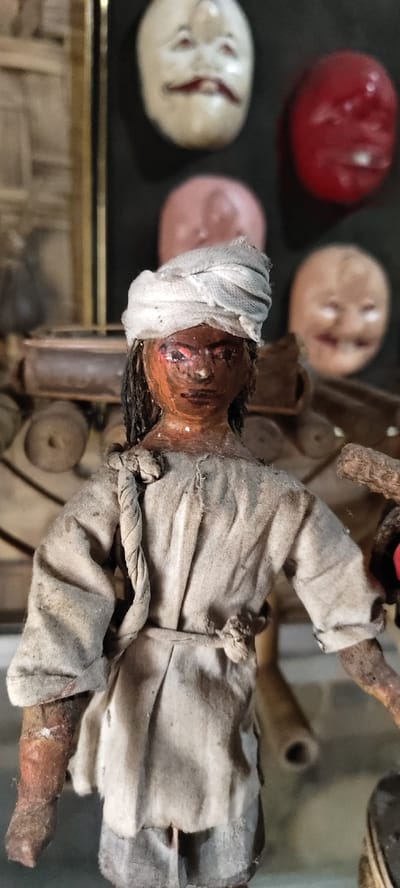A vertical, close-up image captures a weathered, dark-skinned humanoid figurine positioned in the foreground, located within what appears to be a display or workshop setting in Cikanyere, Indonesia. The figurine features a dark brown painted face with red accents around the eyes and dark, thin lips. Its head is adorned with a white, turban-like cloth, and stringy dark material, resembling hair, frames its face. The body is clothed in an aged, off-white or light grey fabric tunic, secured with a thin, rope-like belt, and matching trousers. A thicker, braided rope-like element is draped over its left shoulder, extending down its back. The figurine's limbs appear to be made of a dark, textured material, possibly wood, with visible signs of wear and dust.
The background, out of focus, reveals several other face-like objects or masks against a dark surface. Prominently visible is a cream-colored face with red lips, red eye markings, and a dark, curved feature below the nose. To its right, a solid red, oval-shaped face is present. Partially visible below these are two more faces: one light pink and another light brown/beige, all out of focus. Additional blurry, wooden, or organic-looking objects are discernible behind the main figurine, suggesting a collection or artisan environment. The overall scene indicates a static display of handcrafted items.