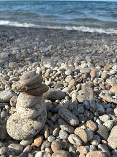 The image shows a stack of smooth, grey and tan stones on a rocky beach. The stones are varied in size and shape, with some appearing flat and others more rounded. They are arranged in a vertical pile, with the largest stone at the bottom. The beach is covered in more of the same types of stones, stretching towards a body of water. In the background, the water is a deep blue, with small waves breaking on the shore. A narrow strip of white foam marks the waterline. The sky above is a pale blue, with some hazy clouds visible. The overall scene is one of natural tranquility, with the focus on the stacked stones. There are no people or animals visible in the image. The scene is located on the shore of Lake Baikal in Tolbazikha, Russia.