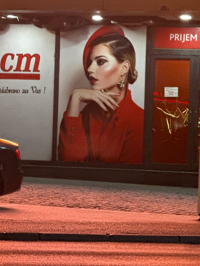 The image shows the exterior of a CM cosmetic market in Ilidža, Bosnia and Herzegovina at night. A large advertisement featuring a woman in a red hat and jacket is prominently displayed on the building. The woman has dramatic makeup and is posing with her hand near her face. To the right of the advertisement is an entrance with a red sign that says "PRIJEM". Through the glass door, reflections of lights and what appears to be some interior fixtures are visible. Below the advertisement, text is partially visible, including "CM COSMETIC MARKET" and "odabrano za Vas!". A dark-colored car is partially visible on the left side of the image. The ground in front of the building is paved with cobblestones, and the scene is illuminated by artificial light, casting a warm glow on the area.