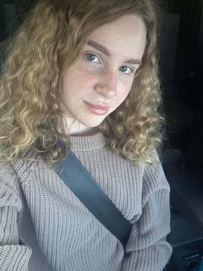 A young, fair-skinned person with curly, light brown hair is wearing a light brown knitted sweater and a dark grey seatbelt in a car. The person is looking at the camera. The background is dark and blurry. The image gives no indication of location in the city of Васильево, Russia.