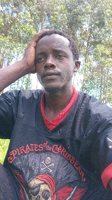 A close-up outdoor portrait depicts an adult male of African descent, identified as being in Gesima ward, Kenya. The individual, appearing to be in his 20s or 30s, possesses dark skin, short dark curly hair with a receding hairline, and sparse stubble on his chin and upper lip, which exhibits a lighter, possibly dusty appearance. His right hand is raised, with fingers resting on his forehead and the right side of his scalp. He is wearing a black short-sleeved jersey-style shirt with a red V-neck collar and red trim on the sleeve ends. The front of the shirt displays a prominent graphic: "PIRATES of the CARIBBEAN" in red stylized text, centered above a white skull and crossbones emblem. Below this is a larger depiction of a skull wearing a red bandana and an eye patch. The subject's gaze is directed slightly upwards and to the right of the camera. The background consists of numerous trees with green foliage, visible against a bright sky, indicating an outdoor, vegetated environment during daylight hours.