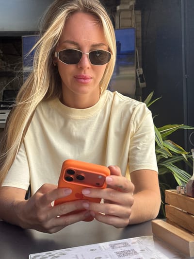 A blonde woman with sunglasses and a light yellow t-shirt is sitting at a table, holding an orange smartphone. She is looking down at the phone, which she is holding with both hands. The background appears to be a cafe or restaurant with plants and wooden elements. The lighting suggests it is daytime. The image was taken in La Vila Joiosa, Spain.