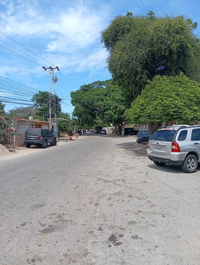 The image depicts a street scene in Atamo Norte, Venezuela. The street is paved and shows signs of wear. On the left side of the road, a dark-colored Jeep is parked near a utility pole with numerous wires attached. On the right, a silver Kia Sportage is parked. Further down the street, several more vehicles are parked along the roadside. The sky is blue with scattered clouds. Trees line the street, providing shade. There are no visible pedestrians or other forms of movement. The location appears to be a residential or suburban neighborhood.