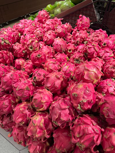 The image presents a vibrant, high-angle view of numerous red-skinned dragon fruits, also known as pitaya, abundantly piled in a retail display. The scene is set in what appears to be the produce section of a grocery store or market in Kelapa Dua, Indonesia.

The dominant subjects are the dragon fruits, which exhibit a striking magenta or fuchsia hue with distinct green-tinged scales. Many of the scales show natural yellowing or slight browning at their edges, indicative of their ripeness and fresh condition. The fruits are loosely stacked, creating a dense, textured mass that fills most of the frame.

To the left, a dark metal grate or shelving structure is partially visible, alongside a glimpse of other green, rounded fruits, possibly avocados or limes, suggesting a varied produce selection nearby. The lighting is uniform, typical of an indoor commercial setting. No people are visible, nor is there any discernible text within the frame. The image focuses on the abundance and visual appeal of the fresh produce.