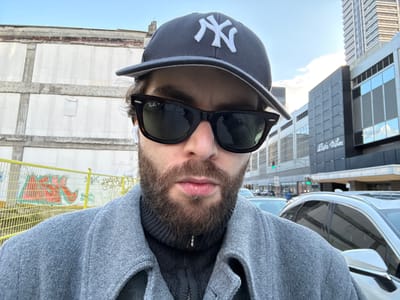 A photograph, oriented upside down, depicts a person wearing a New York Yankees baseball cap and dark sunglasses. The individual has a beard and appears to be wearing a gray jacket. The background shows a cityscape, including a building labeled "Alexis Nihon," with a white vehicle visible in the upper left. A yellow fence is discernible in the background as well. It is not possible to verify that this image shows the city of Westmount, Canada.