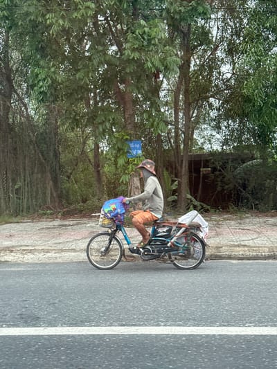 A person is riding a blue bicycle with a basket on the front carrying a colorful bag. The person is wearing a light gray long-sleeved shirt and orange shorts, and a patterned hat. The bicycle also has items strapped to the back. The person is riding on a paved road next to a sidewalk. The background consists of lush green trees and vegetation, with a building visible in the distance. A blue sign with white lettering is mounted on a pole among the trees. The scene appears to be in a rural or suburban area.