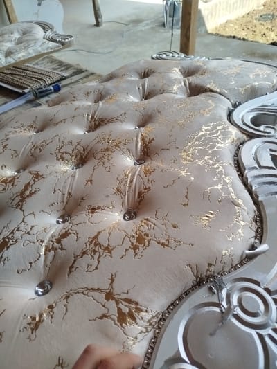 An upholstered, tufted headboard with an ornate silver frame is partially visible. The fabric is light-colored with a metallic gold pattern and has evenly spaced button accents. In the background, another similar headboard and objects are visible. A measuring tape and trim are on the piece. A hand is just visible in the bottom left.