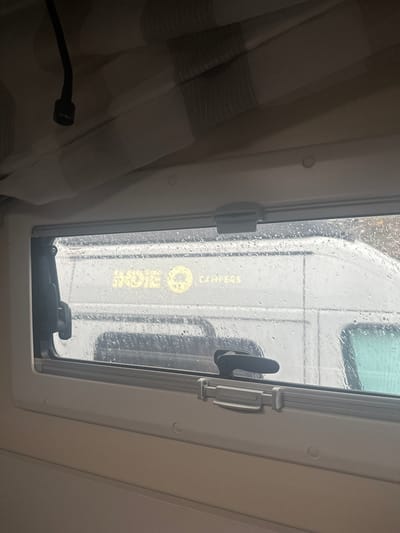 The image shows the interior of a camper van on a rainy day. The view out the window is obscured by raindrops, but the text "ROES CAMPUS" can be seen in yellow on the side of a white vehicle parked outside. A curtain partially covers the left side of the window, and the window's locking mechanism is visible. The location can be inferred to be Montanejos, Spain, based on the text "ROES CAMPUS" seen through the window.