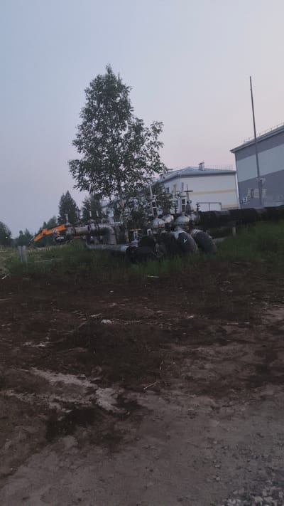 An exterior, eye-level shot shows a construction or industrial site in Kogalym, Russia. In the foreground, dark brown soil and gravel are visible. A patch of green grass and weeds runs through the middle ground. Behind this, a pile of large, black, cylindrical objects, possibly tires or concrete segments, is situated next to an array of large metal pipes and valves. To the left of these pipes, an orange and black JCB excavator is parked. In the background, several trees, some with dense foliage, are present. Further back, a light-colored building with a blue roof and several windows is visible, and to its right, a larger, grey and white industrial building with a tall vertical antenna dominates the background. The sky is overcast and a pale blue-grey.