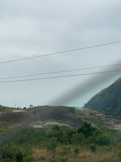 The image shows a view of a rock formation in Kujama, Nigeria, under an overcast sky. Four parallel power lines cut diagonally across the left side of the frame. The rock formation is covered in vegetation, with taller trees and shrubs along the right side and scattered greenery clinging to the rocky surface. A narrow, light-colored band separates the rock formation from the sky.