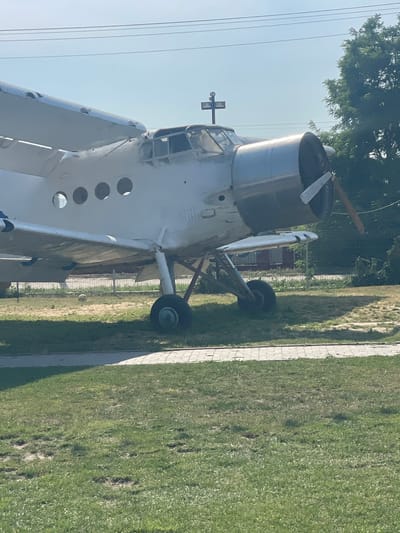 An outdoor, eye-level shot shows a stationary, white Antonov An-2 biplane, facing left. The aircraft is parked on a grassy area adjacent to a paved walkway with a tiled edge. The plane's propeller is visible, as is its engine cowling, cockpit, and part of its wings. A metal fence runs along the background, behind which are trees and a building. A cross-shaped marker is positioned above the cockpit. The sky is clear and blue. The scene is consistent with a static display or a museum exhibit. The location is identified as Zîrnești, Moldova.