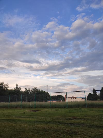 The image depicts an outdoor scene at dusk, likely in the city of Voskobiinyky, Ukraine. In the foreground, a grassy field is visible. A chain-link fence, supported by green metal posts, runs horizontally across the mid-ground. Beyond the fence, a dense line of trees stretches across the landscape. Further in the background, a light-colored building with a blue roof and multiple windows can be seen. To the right of the building, a tall, red cellular tower is situated. The sky above is filled with a mix of blue sky and scattered clouds, with some areas of pink and orange suggesting the setting sun. No people are present in the image.