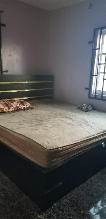 This image depicts an unpopulated indoor bedroom located in Ado-Ekiti, Nigeria. The central subject is a queen-sized bed with a dark-colored wooden frame and a headboard featuring four horizontal gold-colored strips. A light brown or beige mattress, devoid of bedsheets, rests on the bed frame. A single rectangular pillow with a patterned cover (brown, white, and reddish-orange geometric design) is positioned at the head of the bed on the left side. On the right side of the mattress, a small, crumpled textile item is visible. The room's walls are painted a light lavender or pale purple color. A window, fitted with dark-colored metal security bars, is present on the right wall, allowing ambient light into the room. The floor is covered with dark, polished tiles, exhibiting some reflectivity. No individuals are present, and no actions or interactions are depicted.