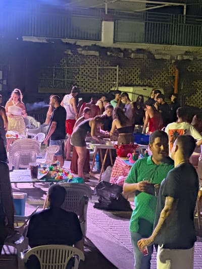 In the city of Blanca, Spain, a group of individuals are gathered at an outdoor event, possibly a party or a gathering. The scene is illuminated by artificial light, casting a purple hue over the surroundings. In the foreground, a man in a black t-shirt is seated, facing away from the camera. To his right, another man wearing a green t-shirt holds a drink, engaged in conversation with a man in a black t-shirt who has visible tattoos on his arm.
Further into the scene, a table is laden with colorful decorations, including what appears to be a miniature volcano surrounded by leis. A red crab figurine and a green cup are also present on the table. Several people are interacting around other tables, some appearing to be preparing food or drinks. A woman in a white top and a patterned skirt stands to the left, holding a drink. Other individuals are standing or sitting in the background, engaged in conversation or observing the activities. The environment suggests a casual social gathering, with plastic chairs and tables set up outdoors. The background features a building with a metal railing and patterned wall.