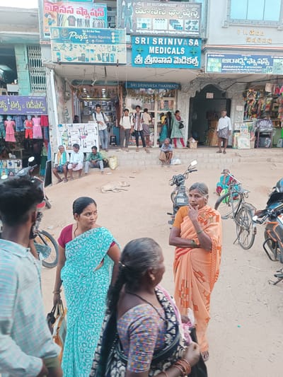 The image depicts a bustling street scene in Kothacheruvu, India, characterized by multiple storefronts and pedestrian activity under daylight.
**Environment:** The scene unfolds on an unpaved, dusty street lined with two-story commercial buildings. Overhead electrical wires are visible across the top of the frame.
**Buildings and Storefronts:**
* **Left:** A building with "MANASAN FASHIONS" and "Pooja Mobile Repairing Centre" signs in Telugu and English, displaying children's clothing on mannequins outside. A closed accordion-style metal gate is visible.
* **Center:** "SRI SRINIVASA MEDICALS" and "SRI SRINIVASA PHARMACY" occupy a building with a prominent green cross symbol and an ornate arched entrance.
* **Right:** "ARADHYA FASHIONS" is indicated by a sign, with various packaged goods and household items displayed outside.
**People:** Approximately 20 individuals are visible, engaged in various activities.
* **Foreground:**
* On the far left, a young man's back is visible, wearing a light checkered shirt.
* In the center-left, a woman in a light blue saree with white patterns holds a dark bag and looks slightly right.
* In the center-right, an older woman in an orange and white patterned saree stands, left hand to her chin, looking right. She wears green bangles.
* On the far right, the back of an older woman with a patterned top and red bangles is partially visible.
* **Midground (on street and steps):**
* A light-colored dog lies on the dusty ground, center-left.
* A woman in a blue saree is seated on the ground on the right, partially obscured by a bicycle.
* On the steps of the leftmost shop, three men are seated: one in a white shirt and green shawl, one in a white shirt, and one in a green shirt.
* On the steps of Sri Srinivasa Pharmacy, several men stand, including one near the entrance in a white shirt and patterned lower garment, and two younger men near a display on the left.
* On the steps of Aradhya Fashions, an older man in a white shirt and dhoti stands, facing right.
* **Background:** Several other individuals are partially visible inside or near the shop entrances.
**Objects:**
* **Vehicles:** A black scooter/moped is parked center-left, a black bicycle stands upright center-right, and the front of an orange and black motorcycle is visible on the far right.
* **Shop Merchandise:** Children's clothes, mobile phone accessories, and various packaged goods are displayed.
* **Other:** Cardboard boxes, a yellow sack, plastic bags, and some litter are scattered on the ground.
**Actions and Interactions:** People are standing, walking, sitting, and conversing, indicating a typical daily street scene with commercial and social activity. The foreground individuals appear to be moving through the space, while others are gathered on the shop steps.