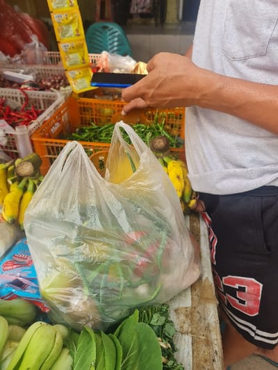 A person sits with groceries displayed, including bananas, green vegetables, and items packaged in a plastic bag. The person holds a blue smartphone next to an orange plastic basket. Other items include yellow packets and produce such as bok choy, all set on a table or countertop. The person wears shorts, one white and the other black with a red and white design.