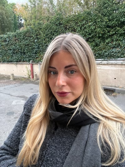 An adult female stands outdoors in Aix-en-Provence, France, depicted from approximately the chest up. The subject, a Caucasian female with long, wavy blonde hair exhibiting darker roots, light-colored eyes, dark, well-defined eyebrows, and dark berry-colored lipstick, gazes directly forward. She wears a dark grey, textured knit sweater and a grey scarf wrapped around her neck, with a darker, possibly black, layer visible beneath. The background consists of a light-colored concrete wall densely covered by green ivy, with taller trees and bamboo stalks visible above, under an overcast sky. On the paved asphalt surface to the left of the subject, two light grey cylindrical bollards and a red vertical pipe are positioned against the wall. A rectangular drain grate is visible on the pavement to her right.