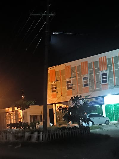 At night in Kotapantonlabu, Indonesia, a view encompasses a multi-story building with patterned facades and windows, a smaller structure with a partial view of its minaret, a white car parked in front, and a picket fence in the foreground. An electrical pole with lines is prominently situated to the left of the buildings. A sign reading "SENTRAL ELEKTRIK" is visible above a storefront. Trees and shrubbery are scattered throughout the scene.