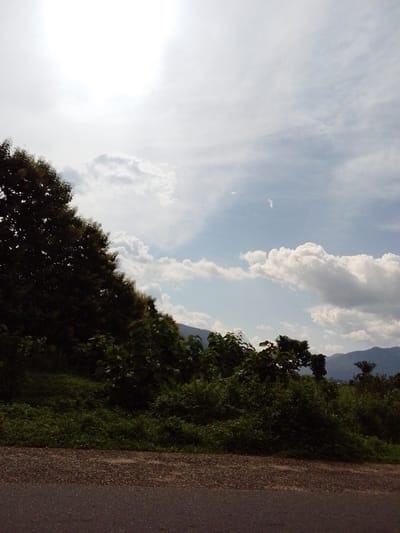 The image depicts a natural landscape scene, likely taken from the side of a road. A paved road, with a rough, gravelly shoulder, occupies the bottom portion of the frame. Beyond the road, a dense, green slope rises, covered with lush vegetation and various trees. The foliage is thick and varied, with some trees being tall and dark, while others are smaller and lighter green. The sky above is a significant element, filled with a mix of bright, hazy sunlight, which appears to be coming from the upper left, and scattered white clouds. In the distance, behind the vegetation, a range of blue-hued mountains is visible, suggesting a location with varied topography. The overall impression is of a rural or semi-rural environment with a bright, partly cloudy sky. The location in Goje, Nigeria, would imply a tropical or subtropical climate, which is consistent with the lush vegetation and the type of trees and mountainous backdrop.