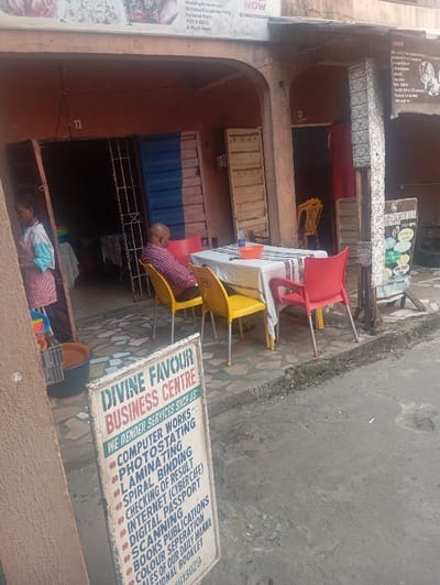 The scene depicts a commercial street in Warri South, Nigeria, featuring the facade of a two-story building with multiple ground-level units. The building is constructed of reddish-brown material, with arched doorways and metal grates or doors. Unit "22" and "23" are labeled above two separate entrances.

In the foreground, a white sandwich board sign for "DIVINE FAVOUR BUSINESS CENTRE" is prominently displayed, listing services such as "COMPUTER WORKS, PHOTOSTATING, LAMINATING, SPIRAL BINDING, CHECKING OF RESULT, INTERNET (CYBER CAFÉ), DIGITAL PASSPORT, SCANNING, BOOKS PUBLICATIONS, COLOUR SEPERATION, SALES OF OUR DAILY MANNA, NATIONAL BOOKLET," and a partial contact number ending in "613029".

Immediately behind the sign, an outdoor seating area is visible. A rectangular table is covered with a white tablecloth and a patterned black and white fabric overlay. A blue plastic bowl containing a clear bottle is centered on the table. Three plastic chairs, two red and one yellow, are positioned around the table. A bald male in a red, white, and blue plaid shirt is seated on a yellow plastic chair at this table, facing away from the camera.

To the left, within an open doorway of unit "22", a person in a light blue and pink striped garment stands near various stacked colorful plastic containers and a gray plastic crate. To the right, another doorway reveals a yellow plastic chair further inside. An upper banner sign on the building advertises "Wedding/Anniversary Birthday/Corporate Party Personal, Party POS & BEDS & Much more." with the text "BOOK NOW 08023333346". Another smaller, darker sign to the right states "WE OFFER THE FOLLOWING..." with partially legible text below.

The ground consists of patterned paving stones directly in front of the building, transitioning to a concrete curb and an asphalt road surface with scattered debris.